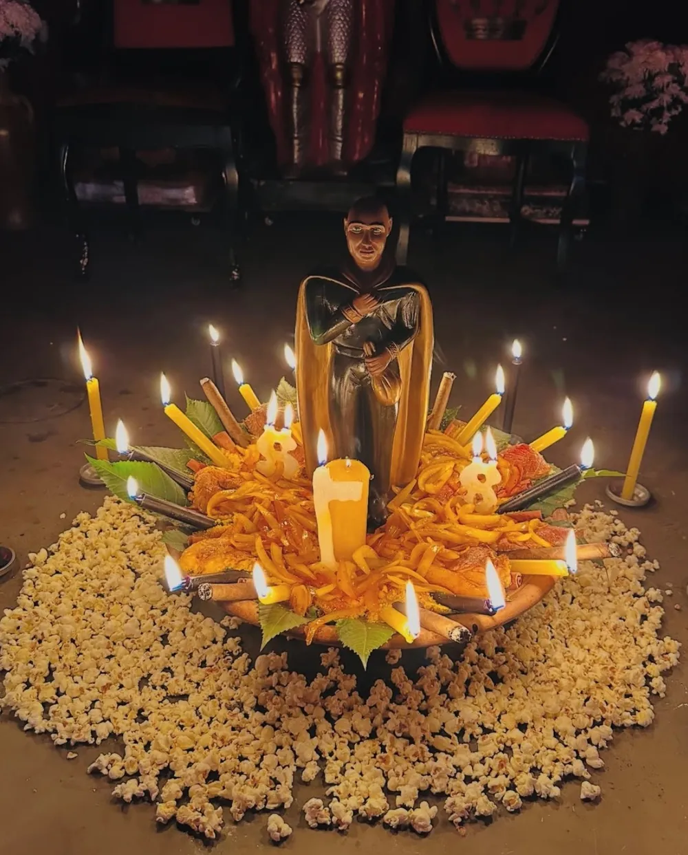 Oferenda com imagem de capa dourada no centro, velas amarelas acesas, charutos e um círculo de pipoca no chão