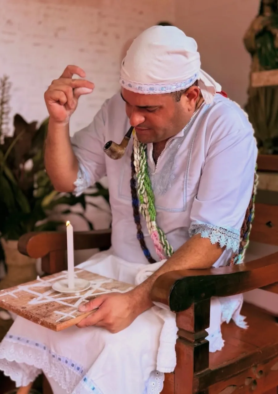 Pai Luiz Fernando de branco, com cachimbo, firmando uma vela sobre uma tábua de ponto riscado em giz, na linha dos Pretos Velhos