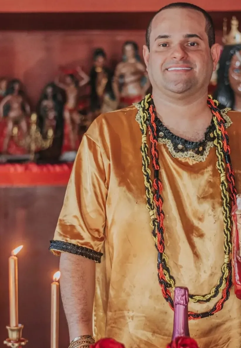 Pai Luiz Fernando de bata dourada e guias vermelhas, pretas e amarelas, sorrindo entre velas acesas e imagens do terreiro